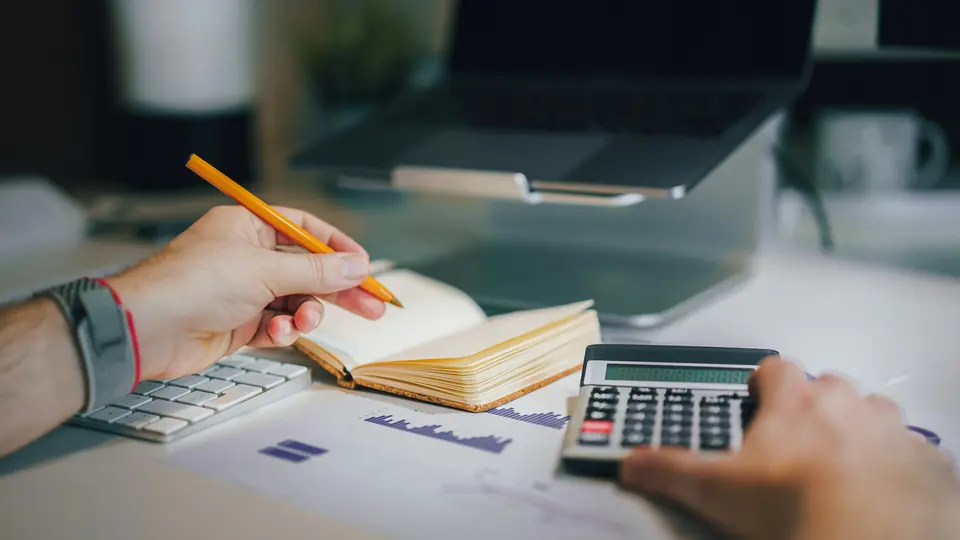 Hands working out a budget with a pencil, a notebook and a calculator, with a laptop behind them