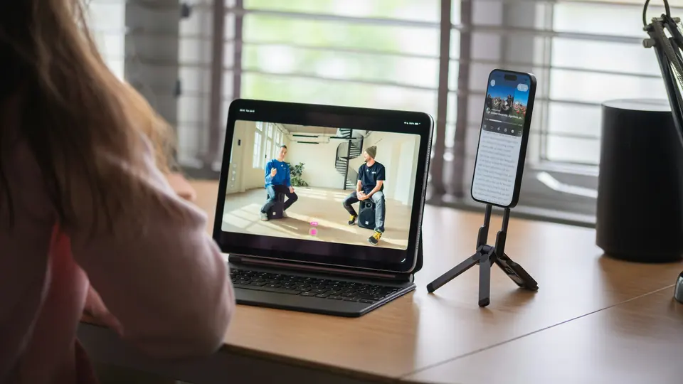 A woman at a desk watching creator videos on a tablet, with a smartphone on a small tripod beside it