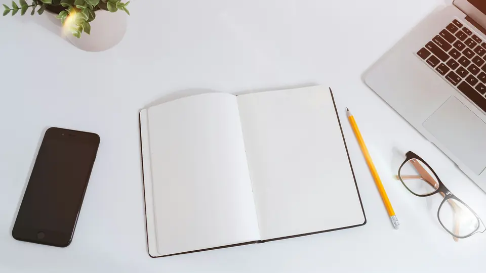 An open blank notebook on a white desk, next to a smartphone, a pencil, glasses and a laptop